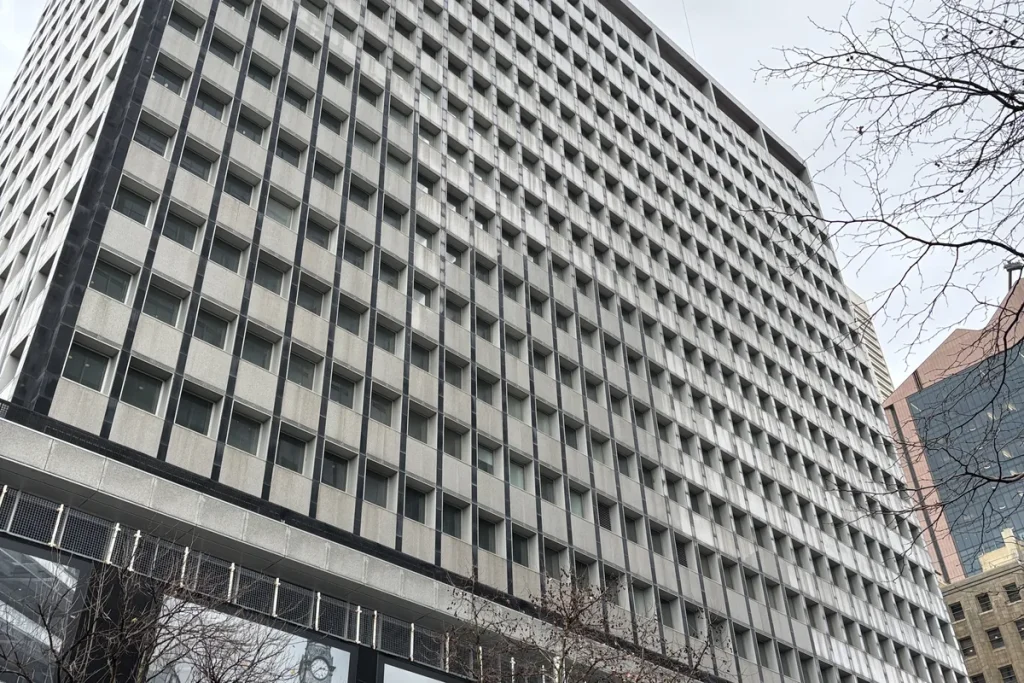 Looking up at the grid of windows on the Reserve Bank of Australia's pale stone head office tower under a grey sky, with bare tree branches at right.
