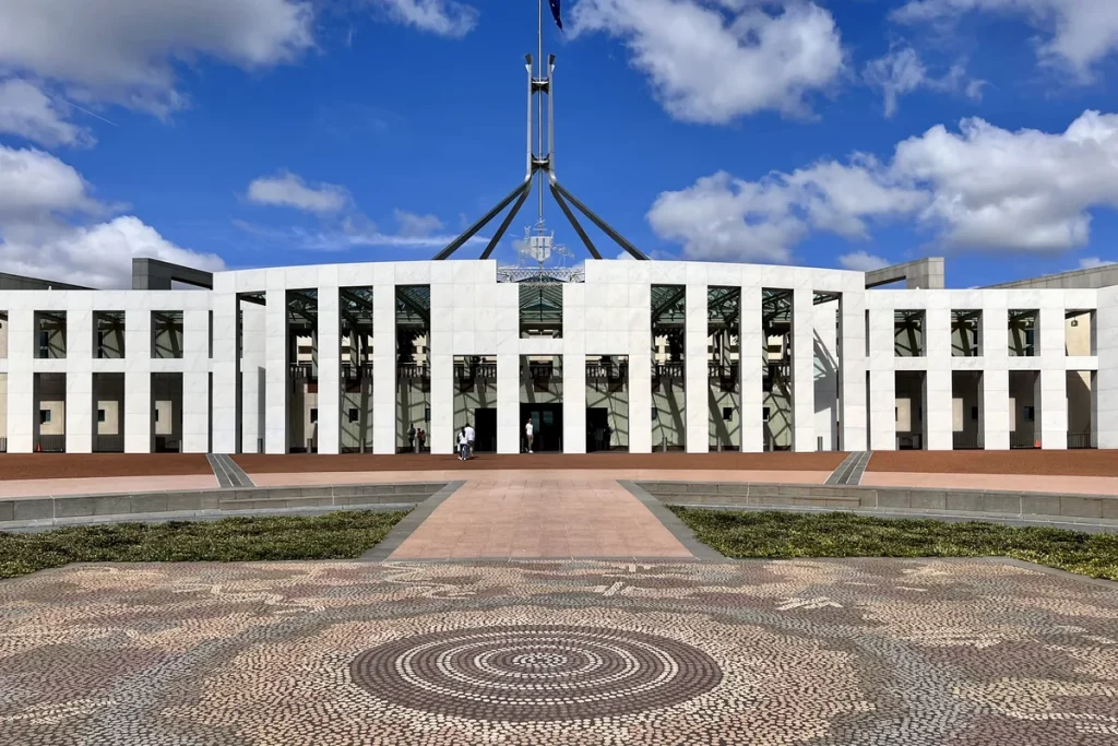 The white colonnaded front of Parliament House in Canberra under a blue sky with puffy clouds, the flagmast above and a circular stone mosaic in the forecourt.