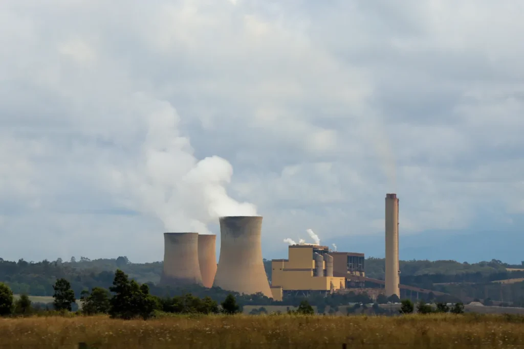 Three cooling towers and a tall chimney of the Yallourn power station venting white steam above dry grassland and a line of trees under a heavy grey sky.