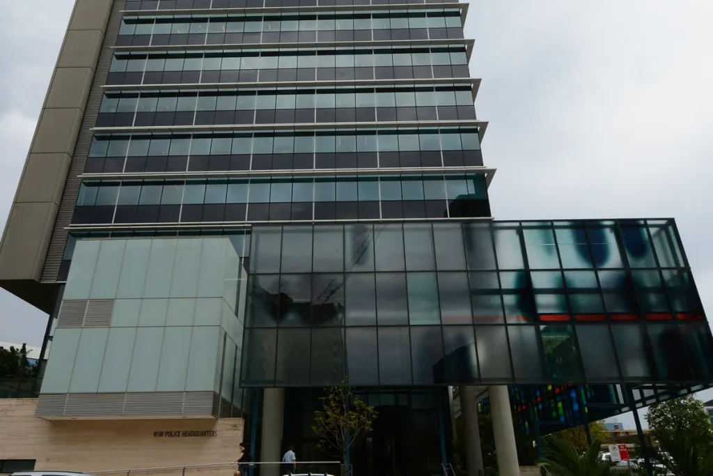 The glass and stone tower of NSW Police Force headquarters in Parramatta under an overcast sky, its name in small letters on the base.