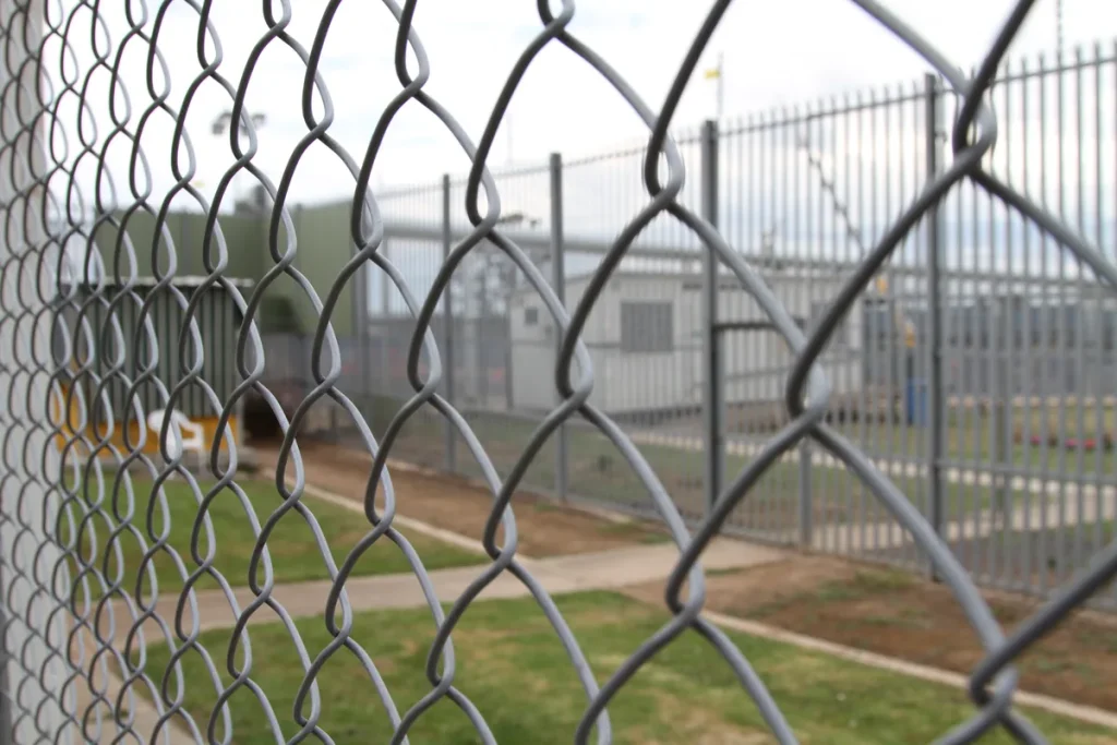 Close chain-link fencing in the foreground with a second high steel palisade fence and low grey buildings blurred behind it.