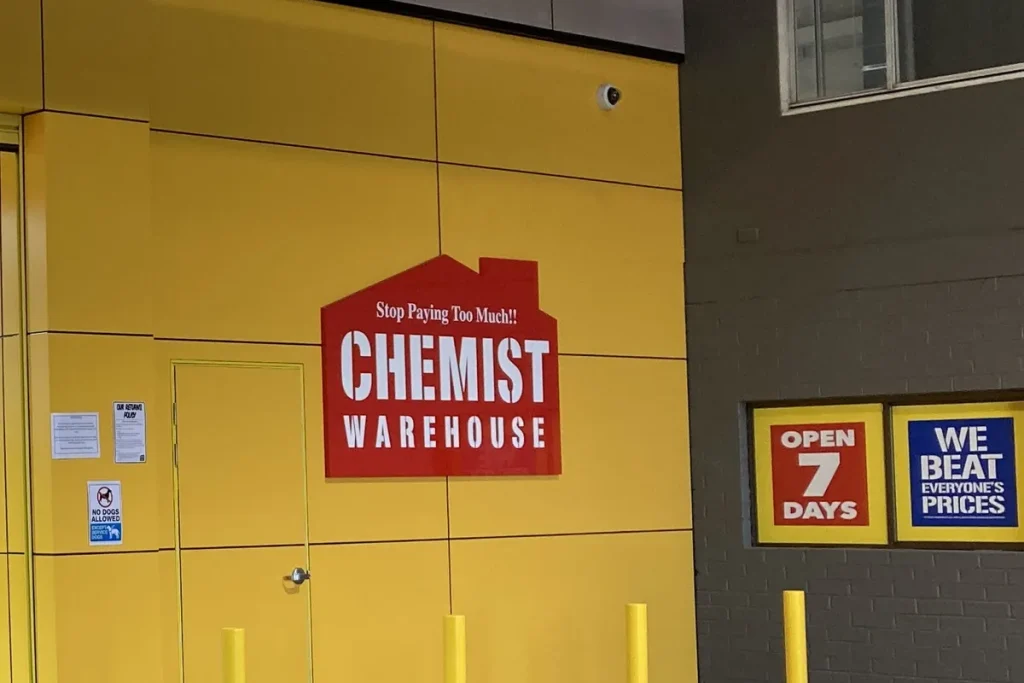 Yellow wall of a Chemist Warehouse store with its red house-shaped logo and window signs reading Open 7 Days and We Beat Everyone's Prices.