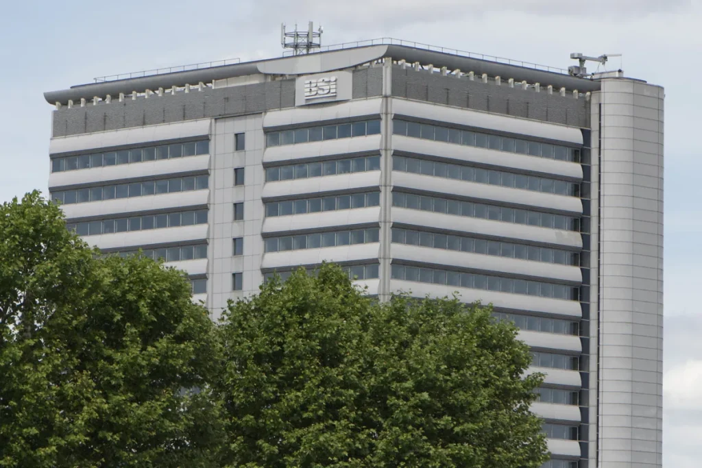 A pale banded office tower with the BSI logo near its roofline, the top floors rising above a line of leafy plane trees.