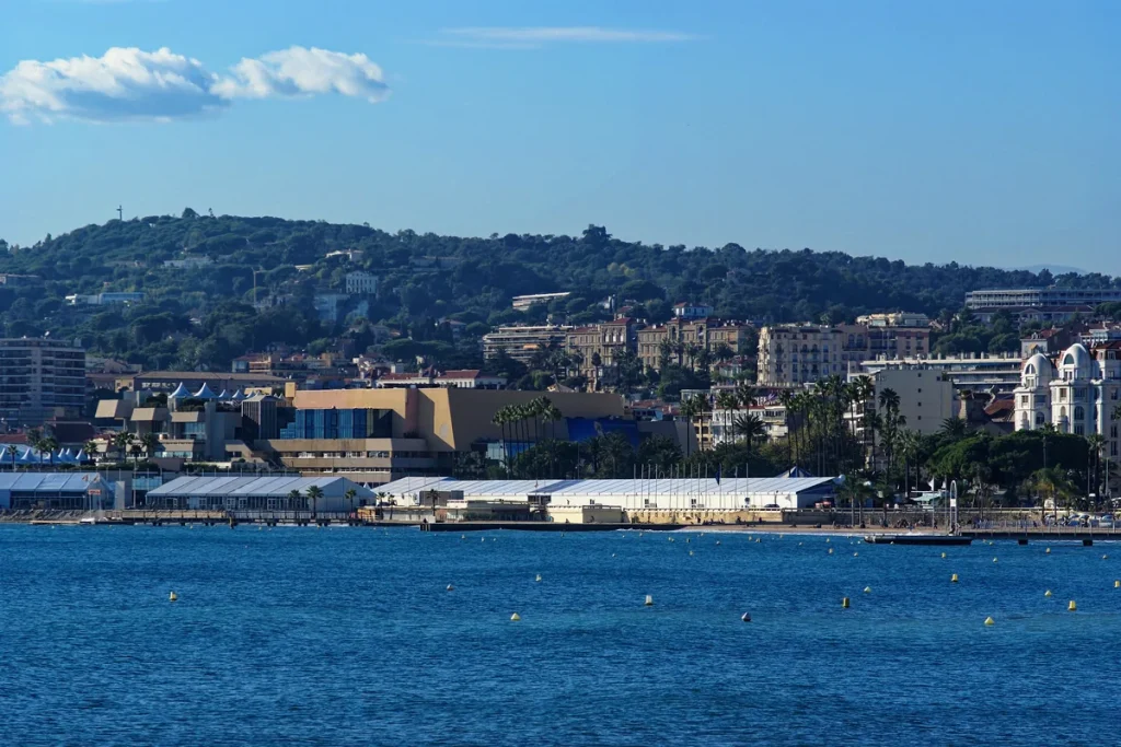 Blue Mediterranean water dotted with yellow buoys in front of the low Palais des Festivals and the white buildings of Cannes, with wooded hills behind.