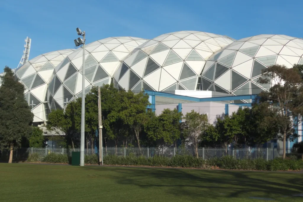 The white geodesic domed roof of Melbourne's rectangular stadium rising behind a row of trees, a light tower and an empty lawn in afternoon sun.
