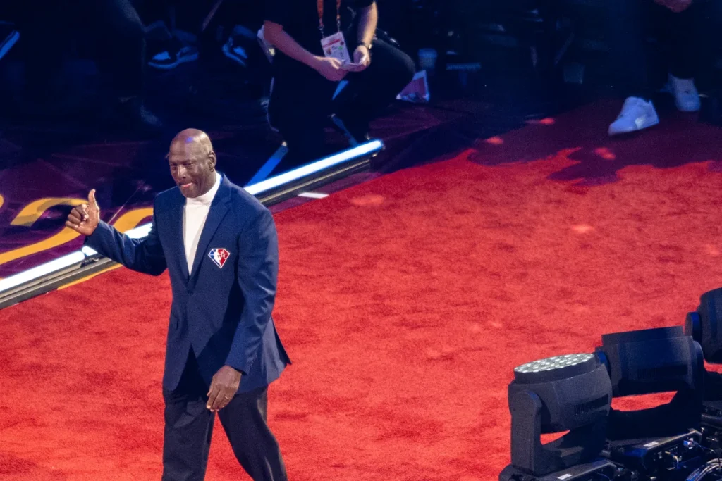 Michael Jordan in a navy blazer and white roll-neck giving a thumbs-up as he walks across a red carpet on an arena floor, stage lights at right.
