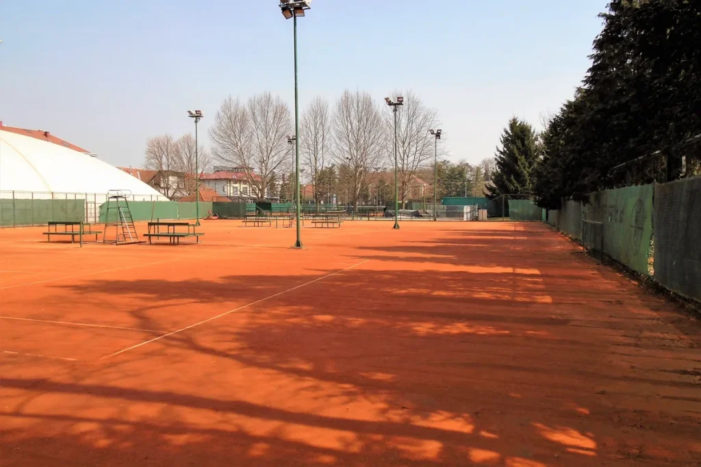Empty orange clay tennis courts in late-winter sun, long tree shadows across the surface, an umpire's chair and benches by the far fence.