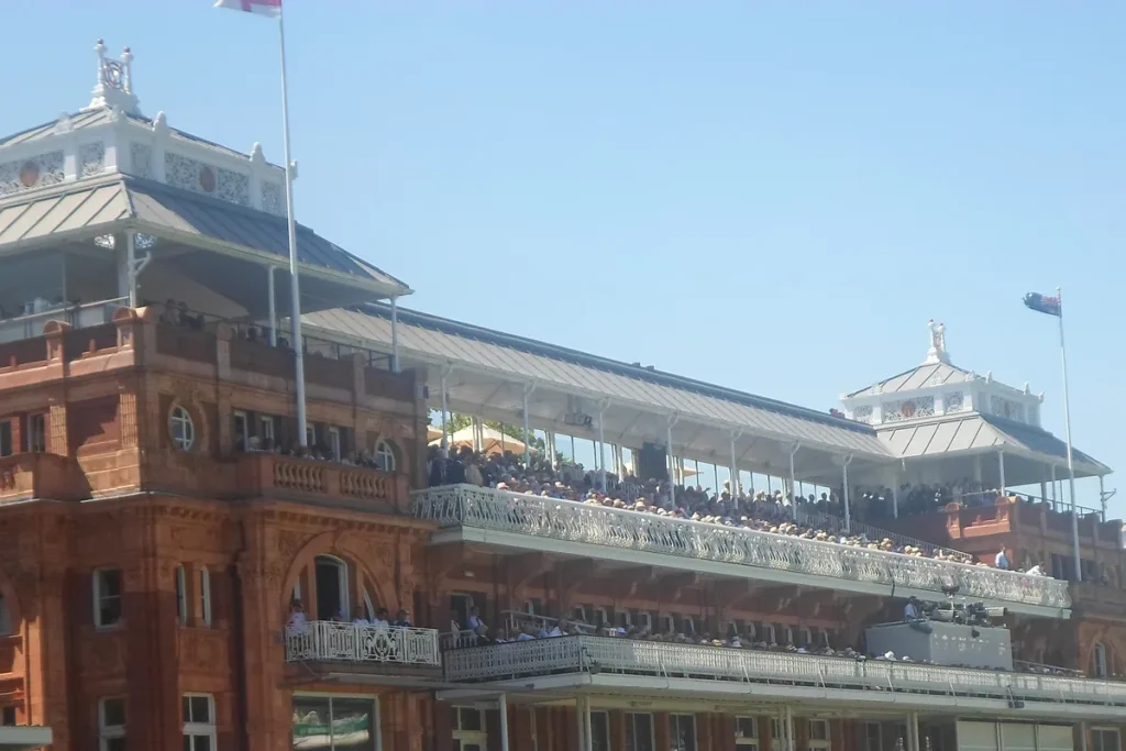 The red-brick Victorian pavilion at Lord's with its white wrought-iron balconies full of spectators and flags flying against a clear sky.