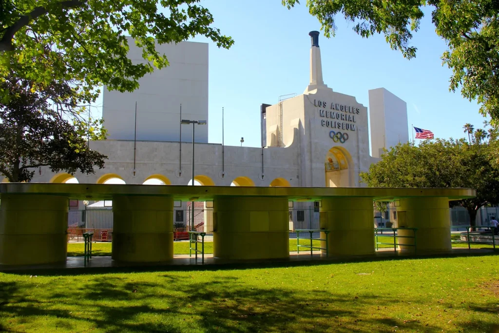 The pale stone peristyle and tower of the Los Angeles Memorial Coliseum with its name and the Olympic rings carved above the arch, framed by trees over a sunlit lawn.