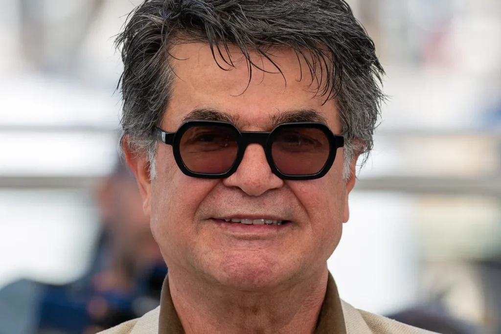 Close portrait of Iranian filmmaker Jafar Panahi, grey-haired and smiling, in dark octagonal sunglasses against a pale blurred seafront.