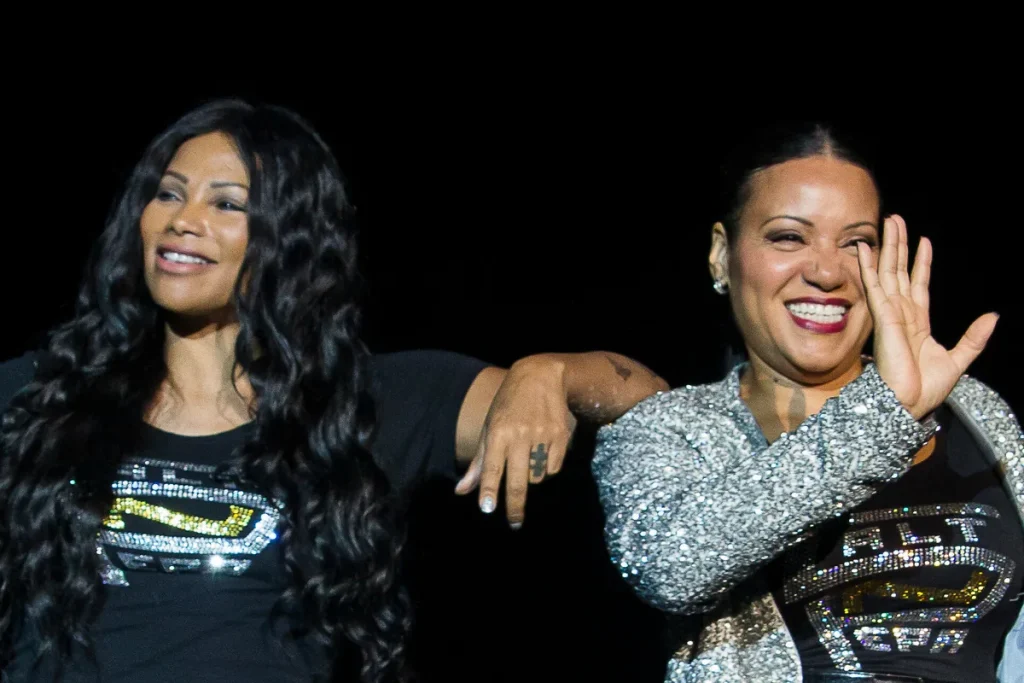 Two women on a dark stage, one with long dark curls and one in a sequinned silver jacket waving, both in black T-shirts with rhinestone lettering.
