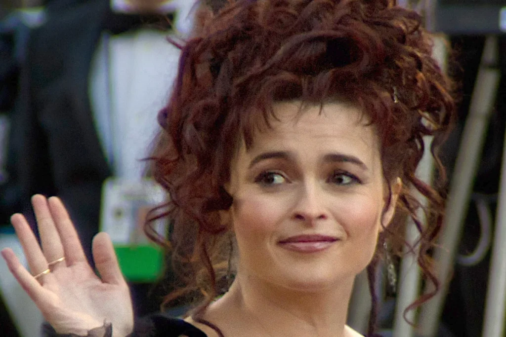 Helena Bonham Carter with piled-up auburn curls, smiling and waving, against a blurred background.