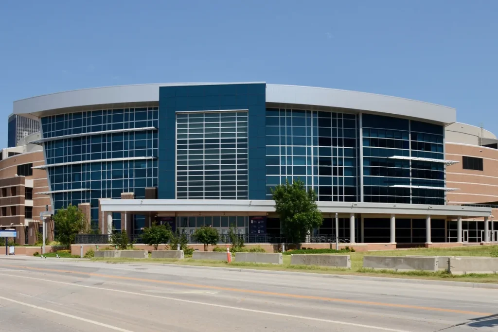 The curved glass and blue-panelled front of Oklahoma City's arena under a clear sky, with concrete barriers and an empty road in the foreground.