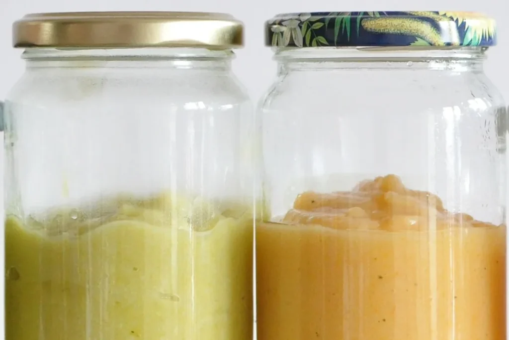 Two glass jars of smooth puree, one pale green and one apricot-coloured, with a gold lid and a patterned lid, against a white wall.