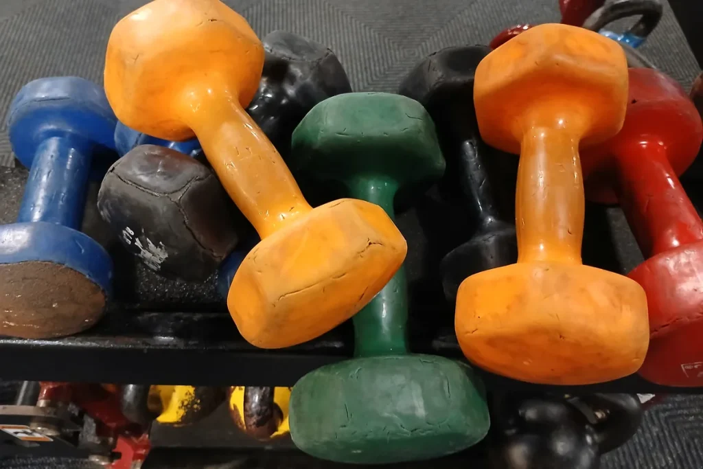 A jumble of chipped orange, green, blue, red and black vinyl dumbbells piled on a gym rack.