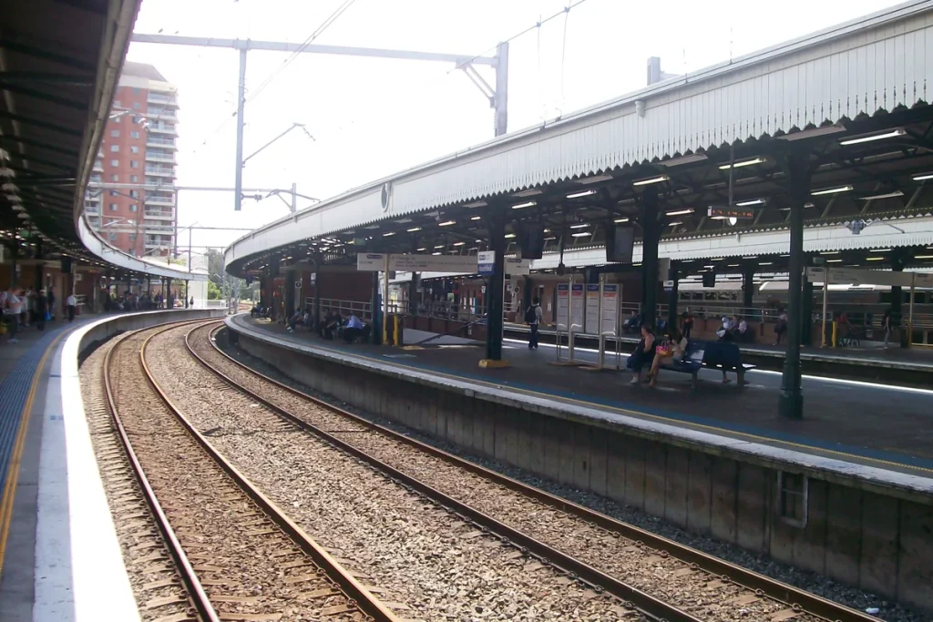 Curving railway tracks and gravel beside a long platform under a scalloped canopy at Strathfield station, overhead wiring gantries against a bright sky and a brick apartment tower behind.