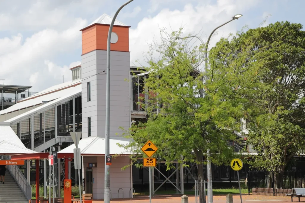 The new St Marys railway station concourse with an orange-topped lift tower, footbridge and leafy trees, under a cloudy sky.