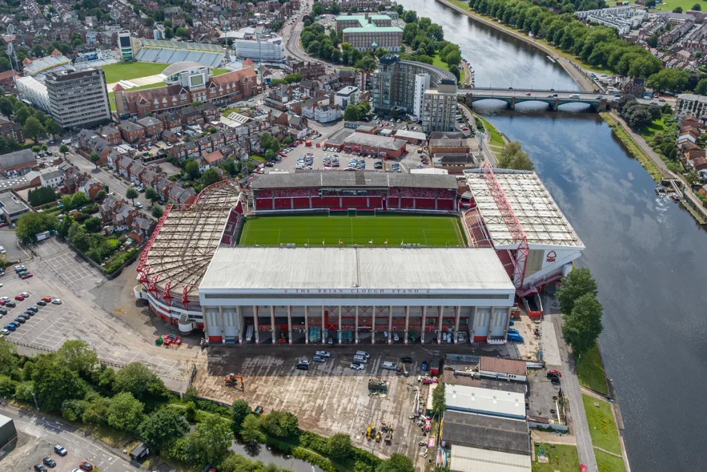 Aerial photograph of a red-seated football stadium on the bank of a wide river, surrounded by terraced houses, with a cricket ground in the distance.