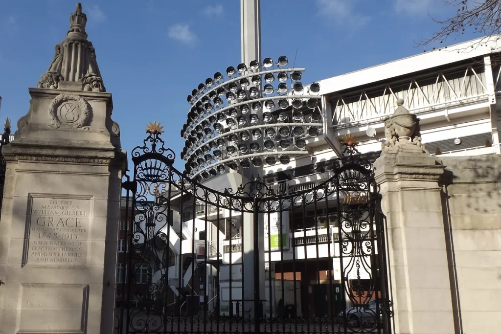 Ornate wrought-iron gates between stone pillars inscribed to the memory of William Gilbert Grace, with a floodlight tower and grandstand behind.