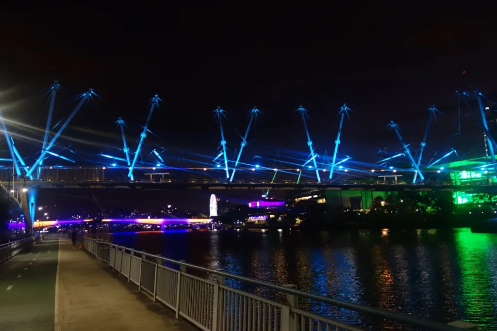 Pedestrian bridge lit in blue across a dark river at night, its spiky mast-and-cable structure reflected in the water beside a riverside path.