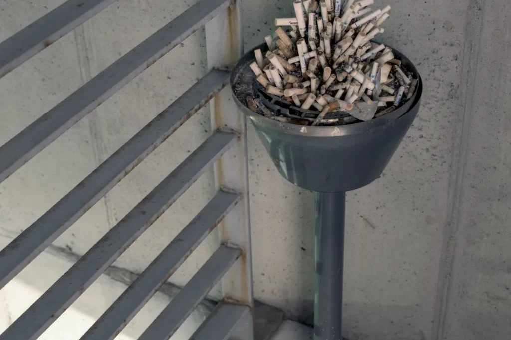 Grey metal standing ashtray crammed with cigarette butts in the corner of a concrete stairwell beside a slatted railing.