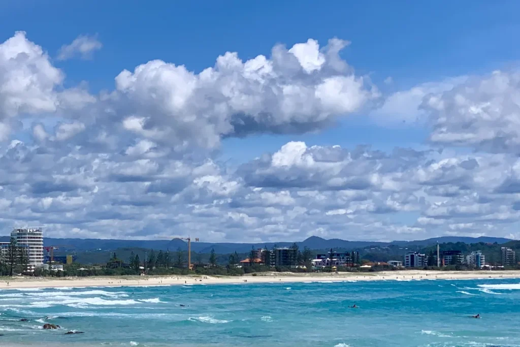 Turquoise surf breaking over dark rocks at Kirra Beach under big white clouds, with a sandy shoreline and Gold Coast high-rises in the distance.