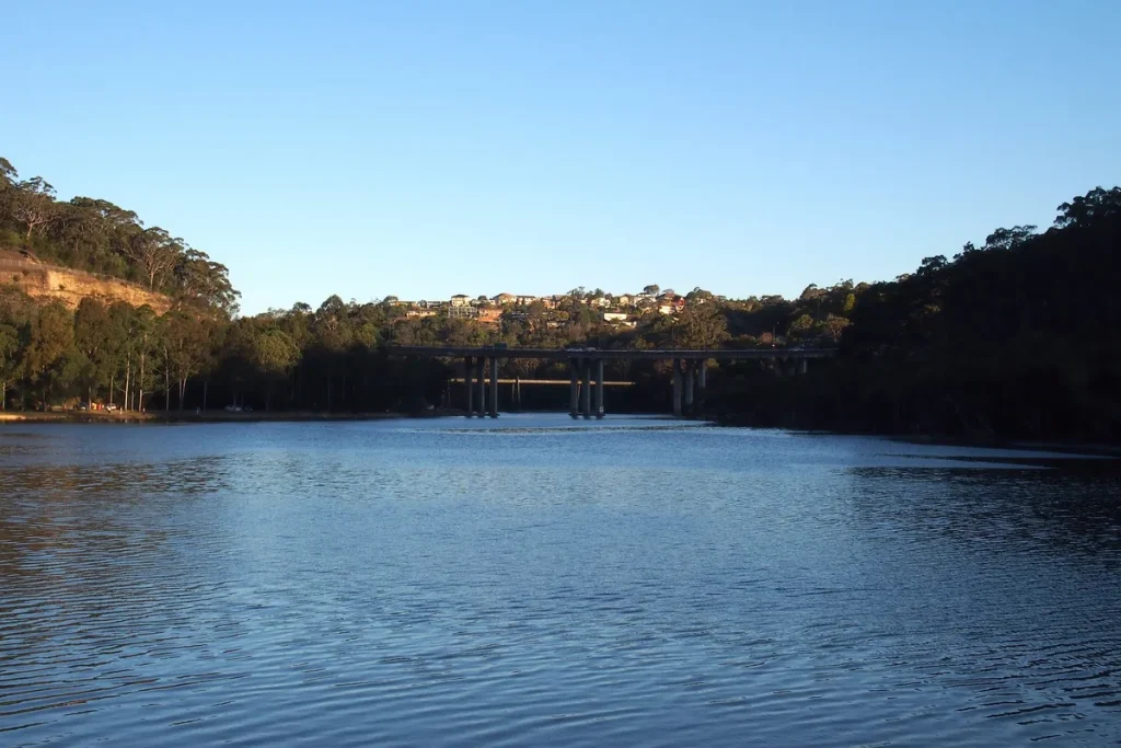 Calm blue water of Middle Harbour stretching to a concrete road bridge, with bushland on both banks and houses on a distant ridge.