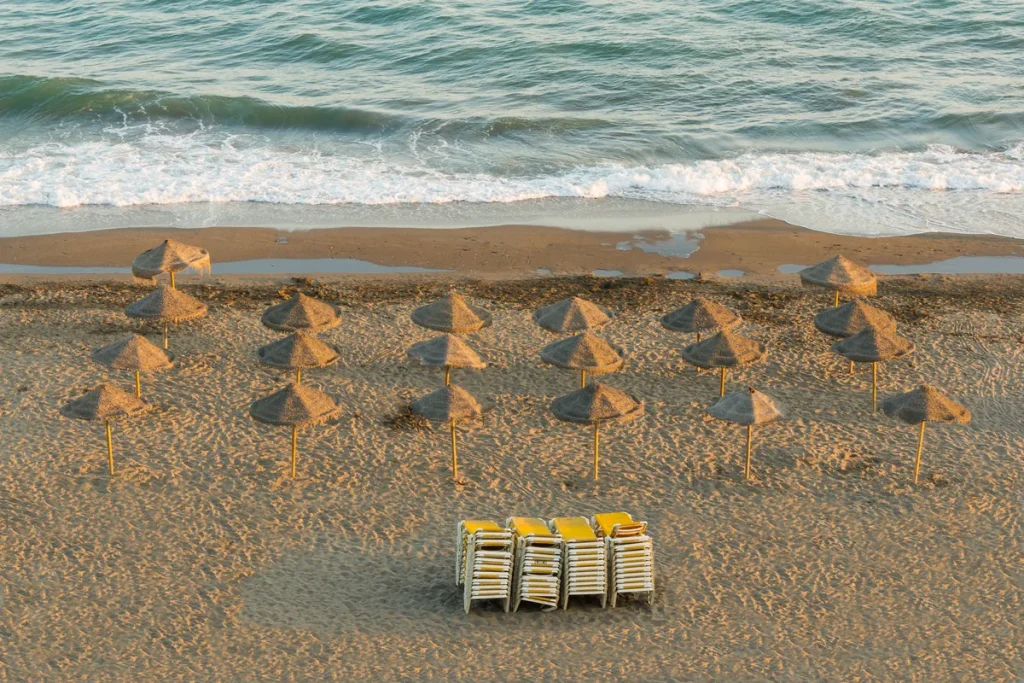 Rows of thatched straw beach parasols on golden sand at the water's edge, with a stack of yellow sun loungers in the foreground and waves behind.