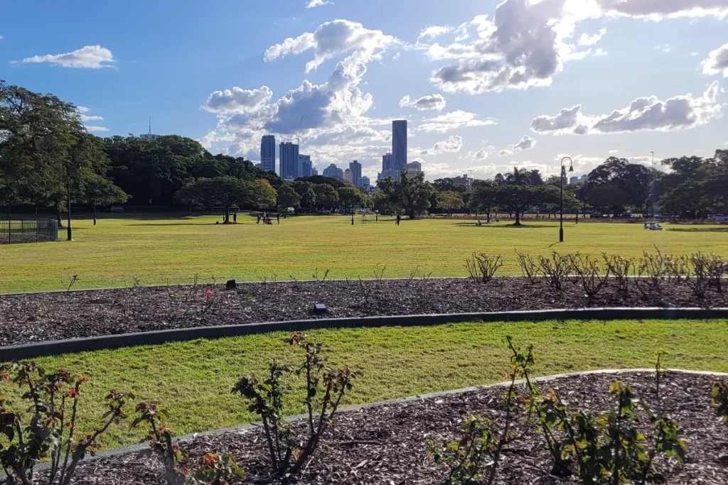 Open green parkland with rose beds in the foreground, shady trees and the Brisbane CBD skyline under scattered cloud.