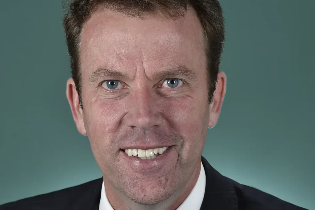 Close head-and-shoulders portrait of Dan Tehan smiling, in a dark suit and white shirt against a teal studio background.