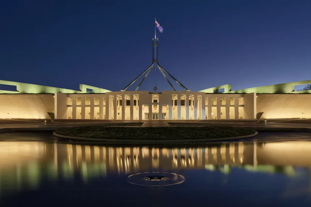 Parliament House in Canberra lit warm gold at dusk, with its flagpole above and the building reflected in a still forecourt pool.