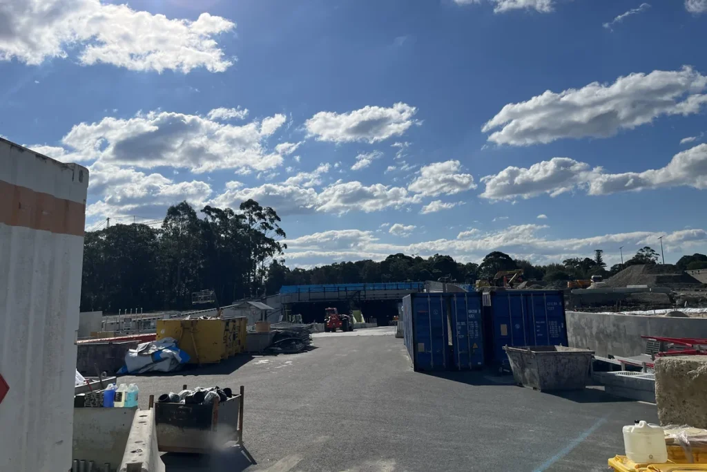 Construction yard with skip bins, shipping containers and a partly built tunnel portal under a bright blue sky with scattered cloud.