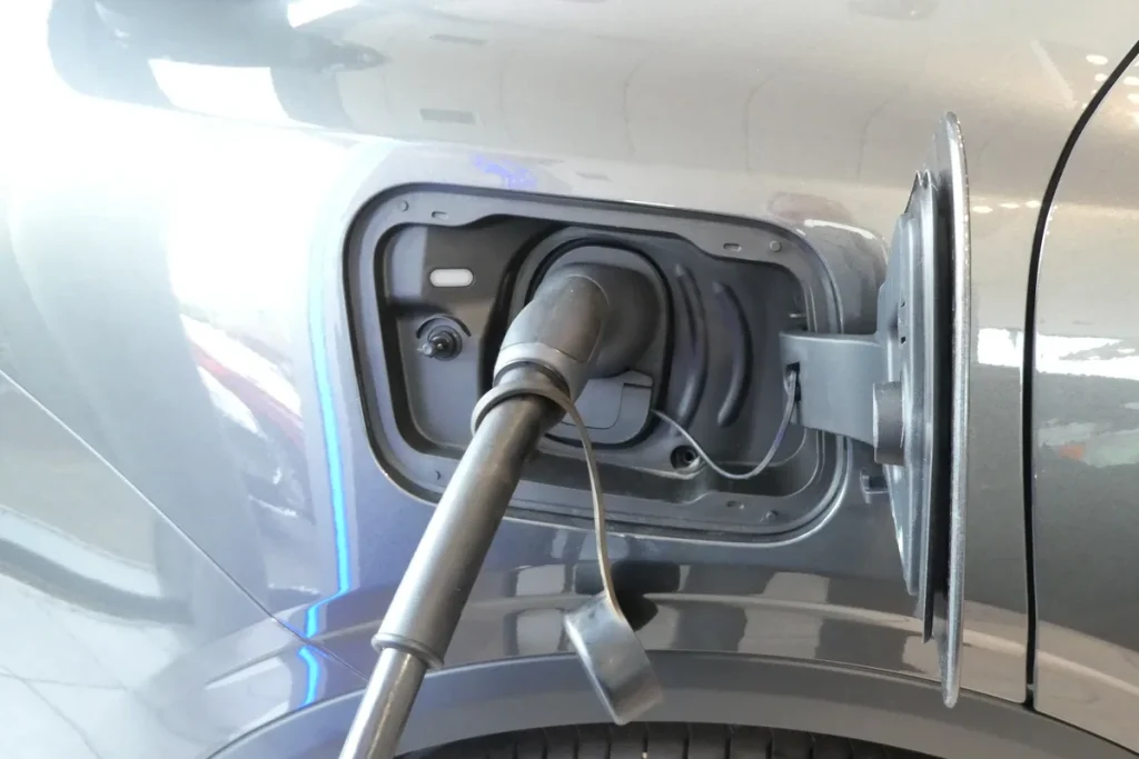 Close-up of a black Type 2 charging plug seated in the open charging port on the silver flank of an electric car, its dust cap hanging below.