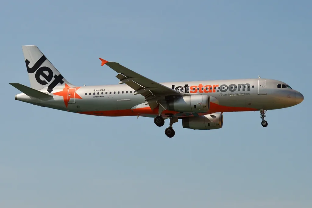 A silver Jetstar Asia Airbus A320 with an orange star and 'Jet' on its tail, wheels down against a pale blue sky.