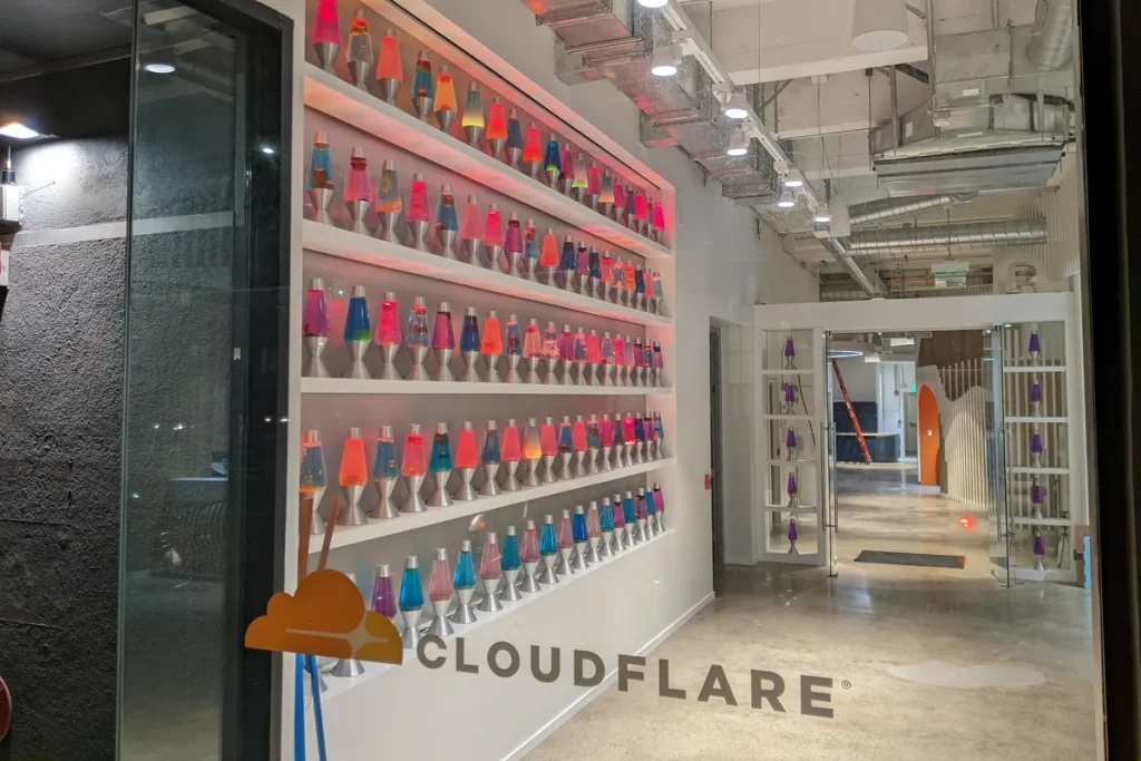 Shelves of colourful lava lamps lining a white lobby wall behind a glass window carrying the Cloudflare name.