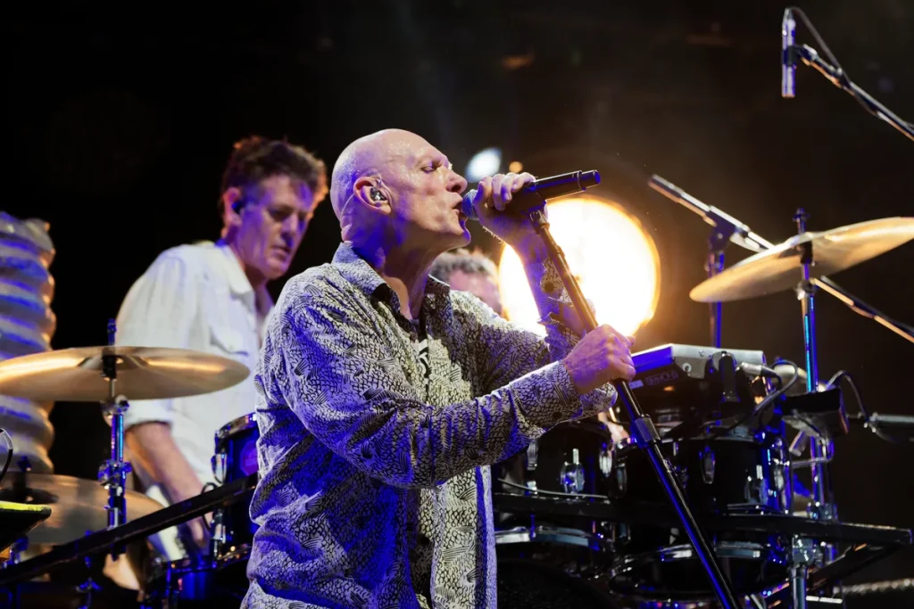 Midnight Oil on stage: a singer in a patterned shirt at the microphone in front, the drummer in a white shirt behind his kit at left, under warm stage lights.