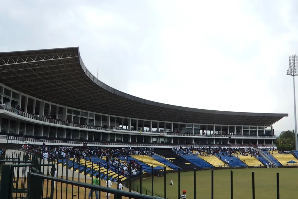 Curved cantilevered grandstand and blue and yellow seating at a cricket ground under an overcast sky, seen through a green railing.
