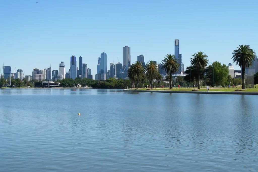 Wide blue lake with palm trees on the far bank and the Melbourne CBD skyline under a clear sky.