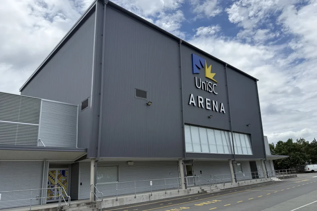 Grey corrugated sports arena with UniSC Arena and the university logo on its facade, under a partly cloudy sky.