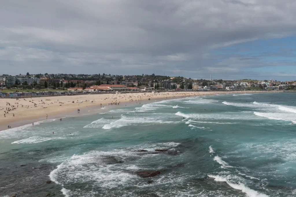 Overcast view along Bondi Beach from the southern headland, surf rolling in over rocks and the Bondi Pavilion on the far foreshore.