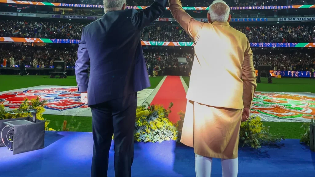 Narendra Modi and Anthony Albanese seen from behind with their arms raised together before a large crowd on a blue stage at Marvel Stadium, Melbourne, at the Melbourne Meets Modi community event on 9 July 2026.