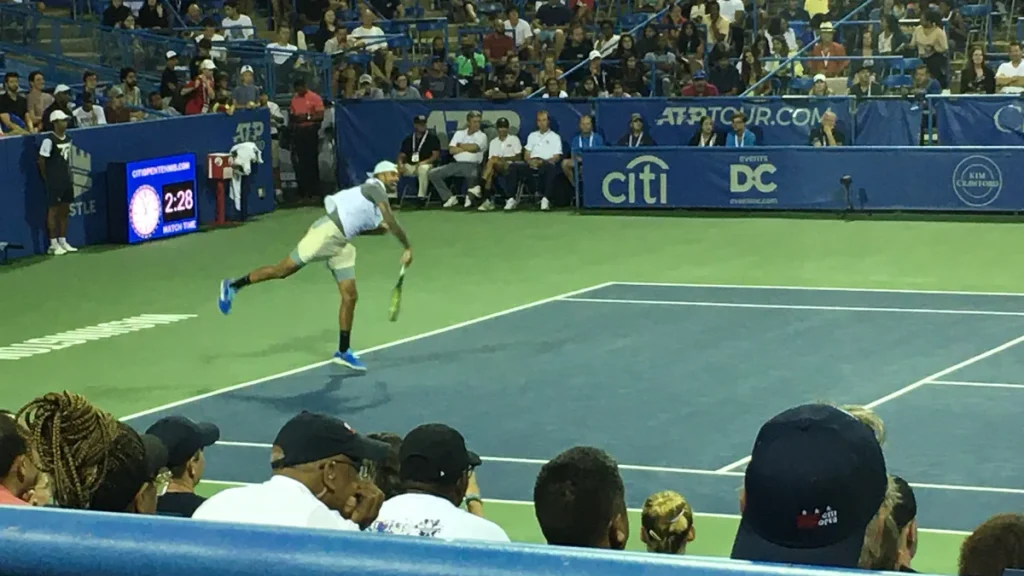 Nick Kyrgios mid-serve on the blue court at the 2022 Washington Open, in a white cap and light kit, seen from the stands with spectators in the foreground and the tournament's boards and match clock beyond; photographed in 2022. From 2022, not from the events in the report.