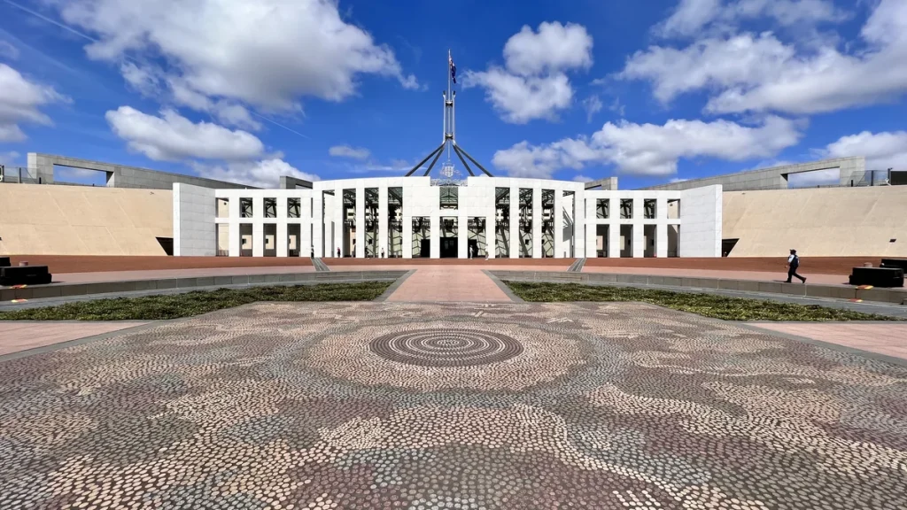 Parliament House in Canberra seen from the front across its mosaic forecourt, with the flag on the central flagpole above the entrance and the building's curved white walls on either side; photographed in March 2022. The building's exterior, not a scene from the Senate vote of 18 August 2026.