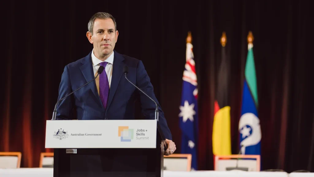 Treasurer Jim Chalmers speaking at a lectern carrying the Australian Government coat of arms and the Jobs and Skills Summit logo, with the Australian, Aboriginal and Torres Strait Islander flags behind him at Parliament House, Canberra; photographed on 2 September 2022. From 2022, not from the report's release.