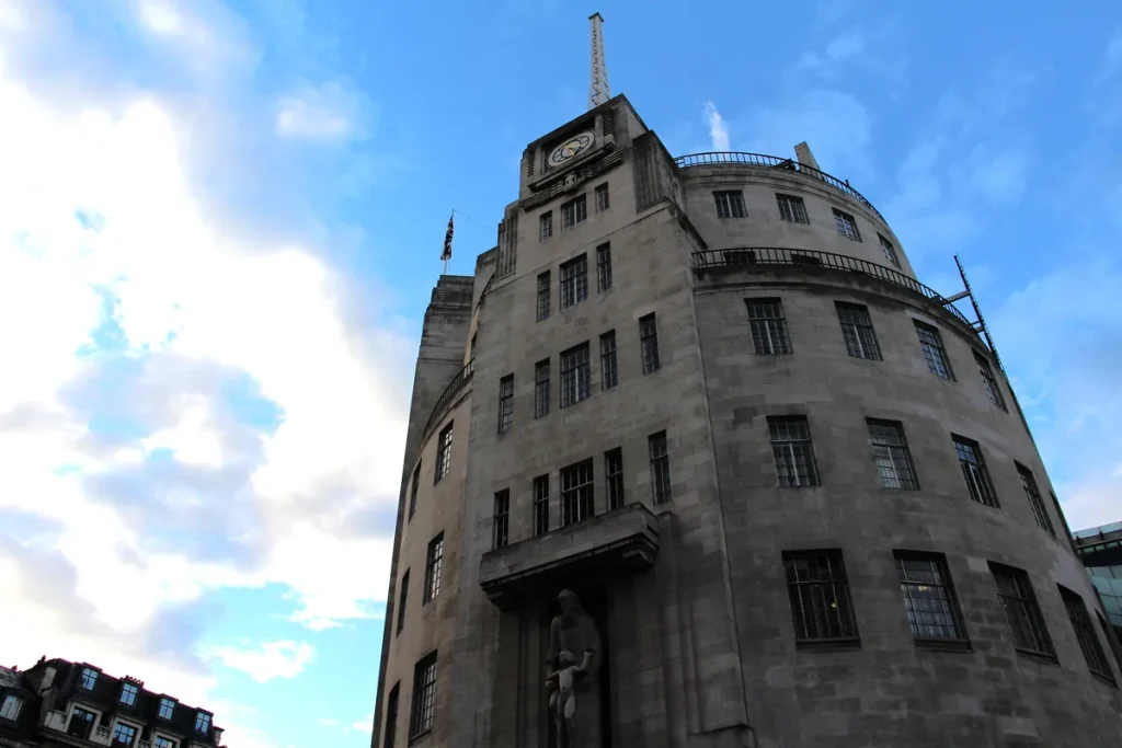 The curved Portland stone facade of BBC Broadcasting House in London rising against a blue sky with white cloud, a mast and clock at the top and a carved statue above the entrance.