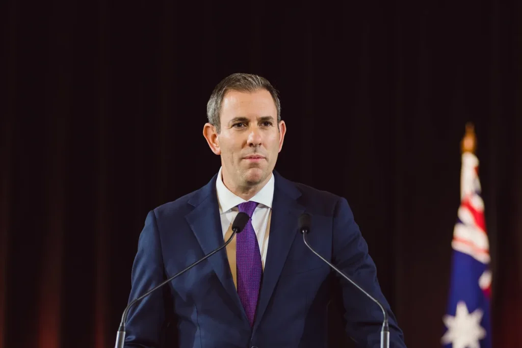 Treasurer Jim Chalmers in a navy suit and purple tie standing behind two microphones, with an Australian flag behind him on the right against a dark curtain.