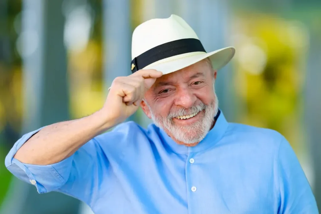 Brazilian President Luiz Inácio Lula da Silva smiling and touching the brim of a white panama hat, wearing a light blue shirt, against a softly blurred green and yellow background.