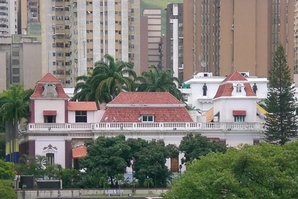 The terracotta-roofed Miraflores Palace in Caracas, with palm trees, two rooftop statues and a Venezuelan flag at left, in front of tall apartment towers.