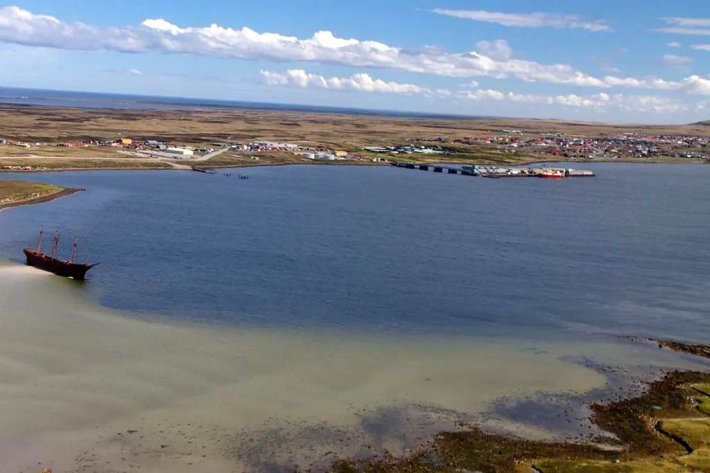 Elevated view across Stanley Harbour in the Falkland Islands, with a rusting three-masted hulk in the shallows and the town and port on the far shore.