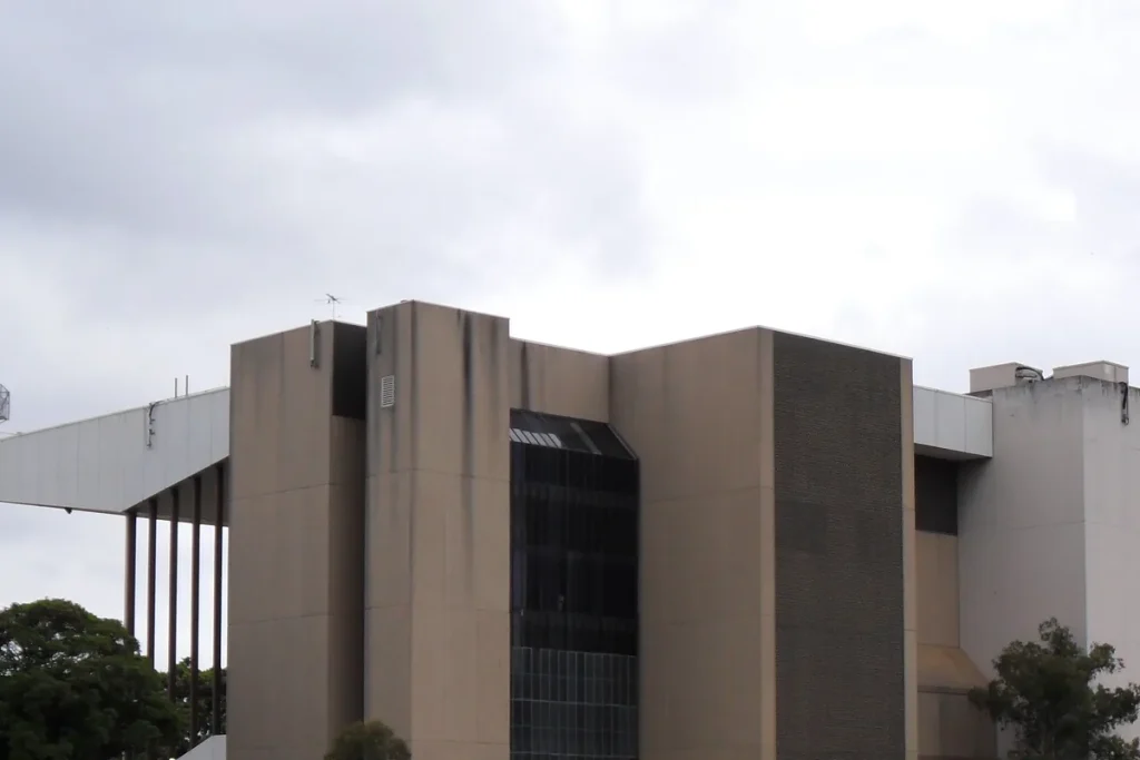 The concrete grandstand at Wentworth Park in Glebe, with tall tan towers, a dark glazed window strip and a cantilevered roof on slender columns, under an overcast sky with trees at its base.